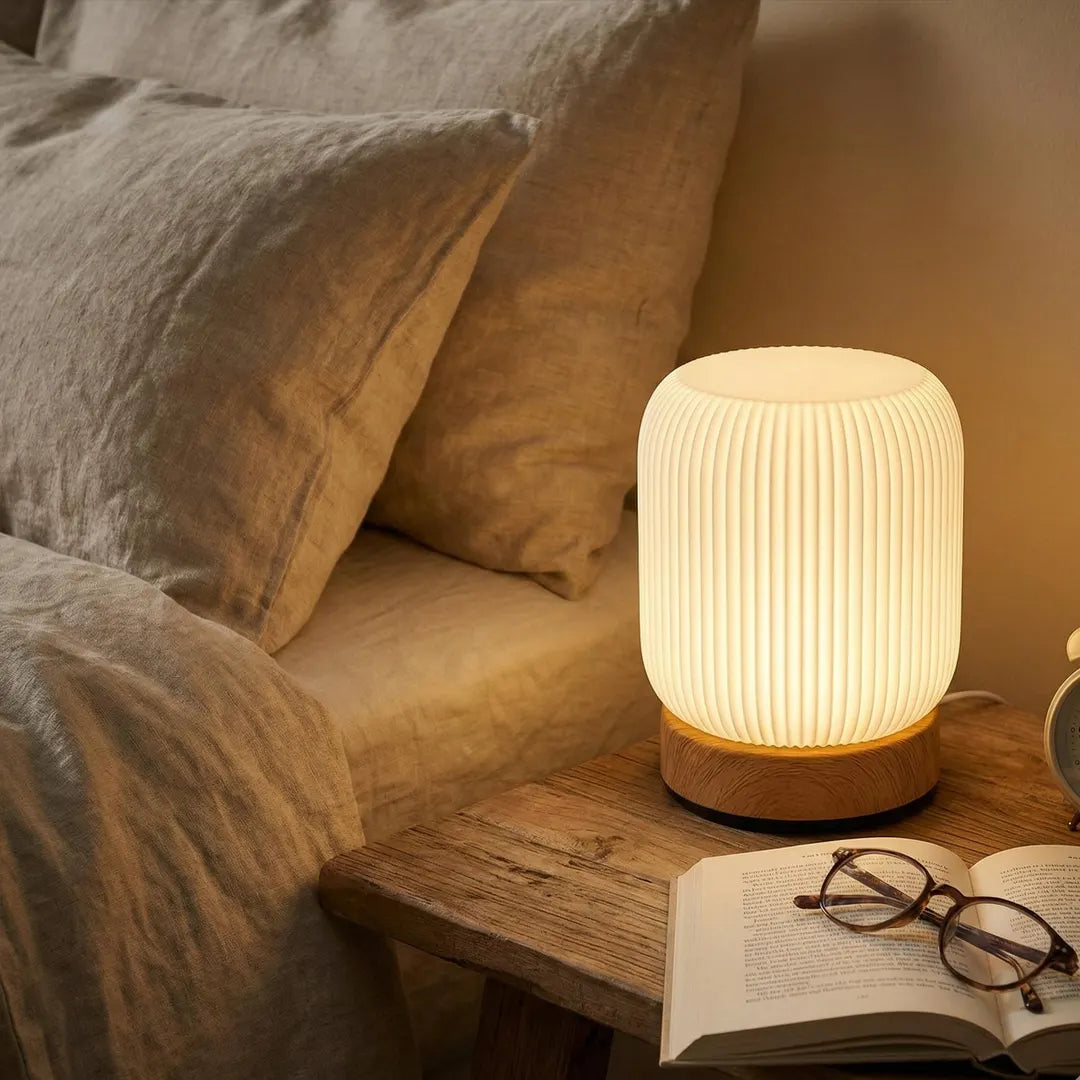 Table lamp on a wooden side table next to an open book and glasses, with pillows in the background